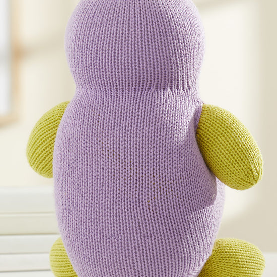 A knitted plush toy with a purple body, yellow arms, legs, and top tuft, sitting on a white surface, facing away from the camera.