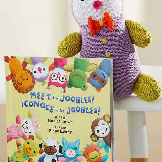 A plush toy in a purple outfit sits on a chair next to a bilingual children‚Äôs book titled Meet the Joobles! ¬°Conoce a los Joobles! featuring colorful animal characters on the cover.