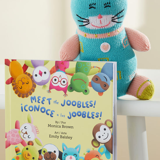 A knitted blue cat plush sits behind a bilingual children‚Äôs book titled Meet the Joobles! ¬°Conoce a los Joobles! on a white chair.
