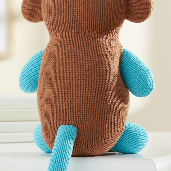 A knitted stuffed monkey toy with a brown body and blue arms, legs, and tail sits on a white surface, facing away.