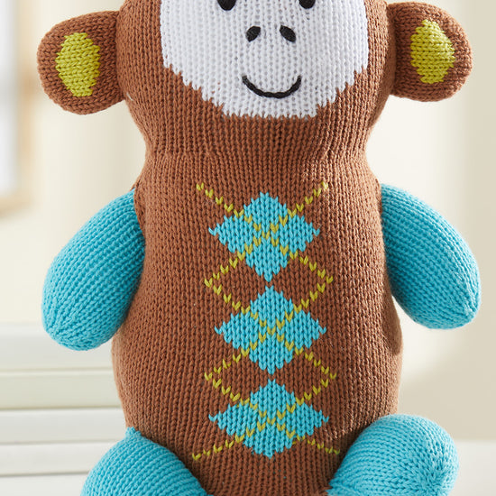 A knitted stuffed monkey with a brown body, blue arms and legs, yellow and green diamond pattern on its chest, and a white face with a smiling expression.