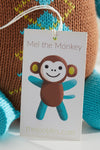 A product tag with an illustration of a smiling cartoon monkey labeled Mel the Monkey and the website thejoobles.com in front of a knitted toy.