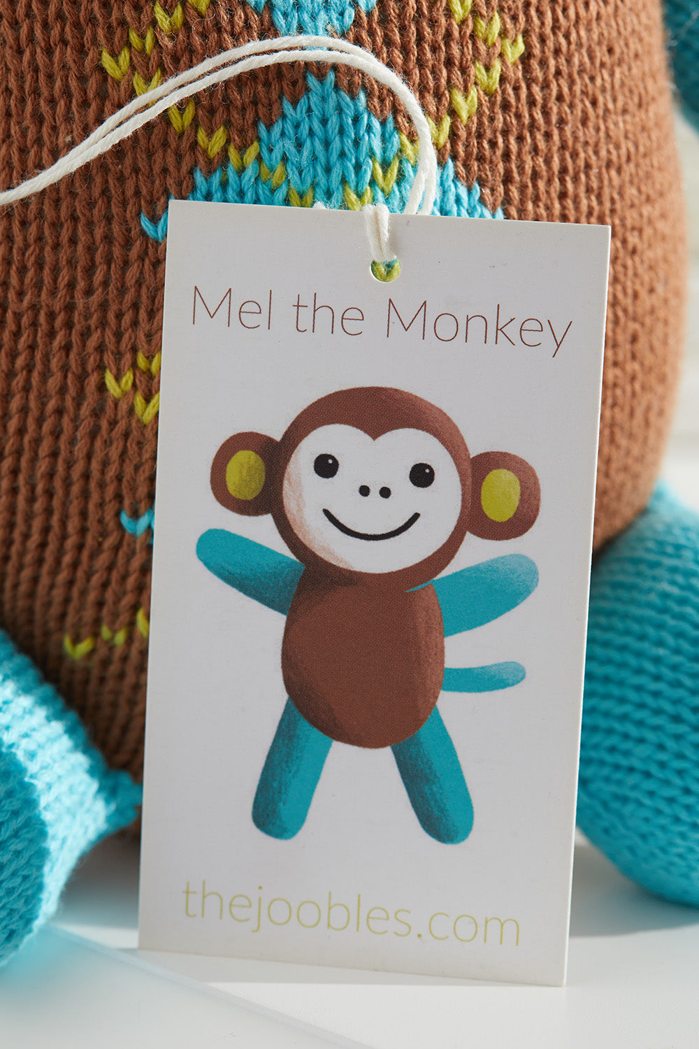 A product tag with an illustration of a smiling cartoon monkey labeled Mel the Monkey and the website thejoobles.com in front of a knitted toy.