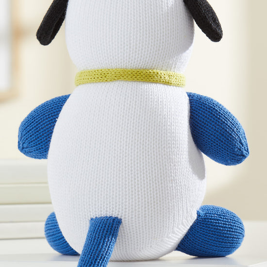 A knitted stuffed dog with a white body, blue legs and tail, black ears, and a yellow collar, sitting on a white surface facing away.