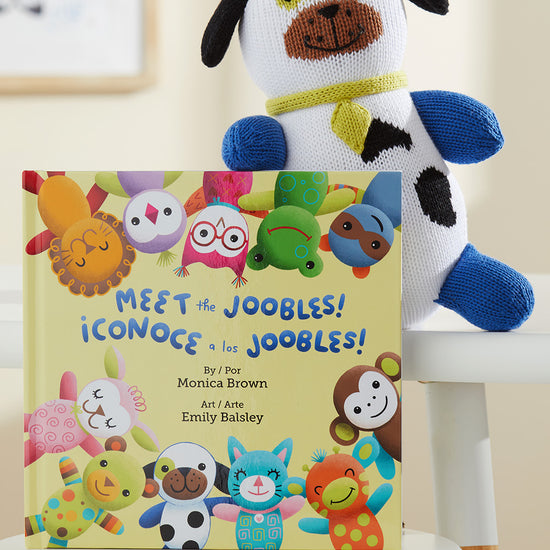 A bilingual childrens book titled Meet the Joobles! ¬°Conoce a los Joobles! is propped on a white chair next to a knit plush dog.