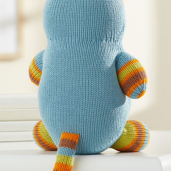 A knitted blue plush toy with striped orange, yellow, and green arms, legs, ears, and tail, shown from the back, sits on a white surface.