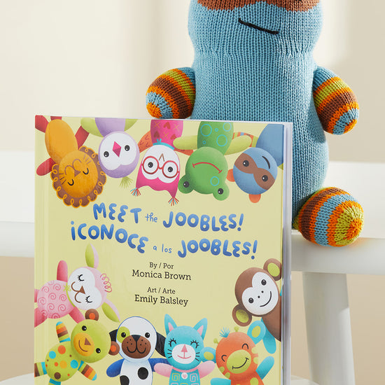 A knitted blue and orange stuffed animal sits behind a bilingual childrens book titled Meet the Joobles! ¬°Conoce a los Joobles! displayed upright on a white chair.