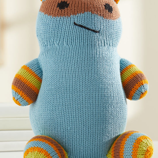 A knitted stuffed toy with blue body, orange and yellow striped limbs, orange ears, and a brown mask-like pattern on its face sits on a white chair.