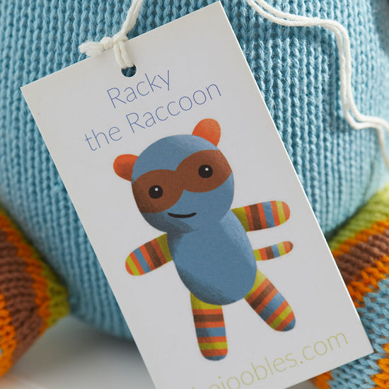 A tag attached to a blue knit toy reads ‚ÄúRacky the Raccoon‚Äù and shows an illustration of a raccoon with striped arms and legs. Thejoobles.com is printed at the bottom.