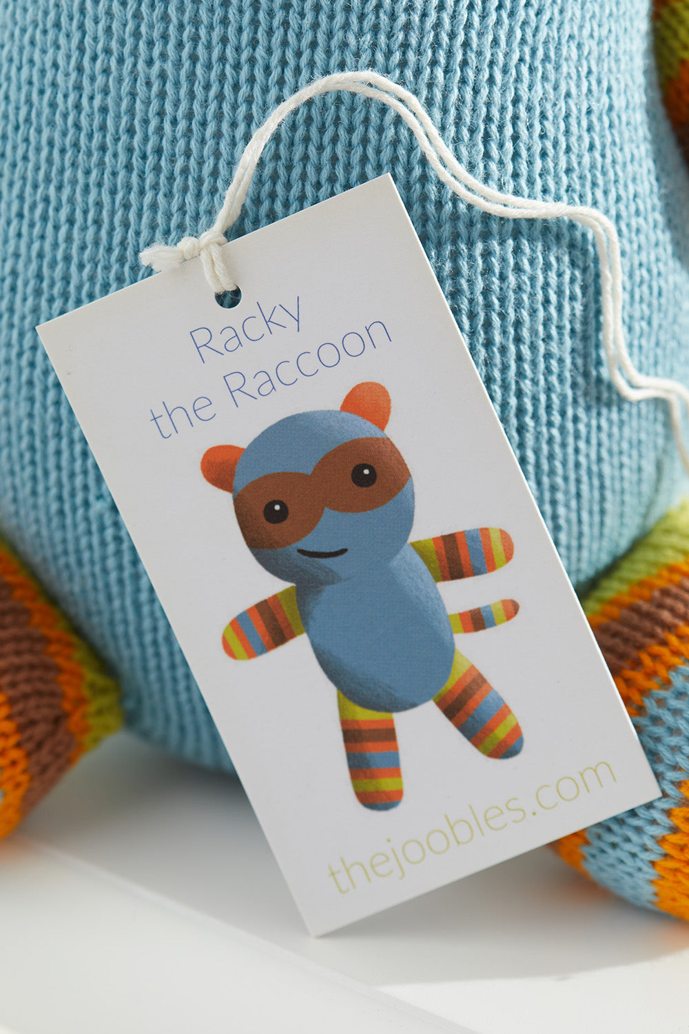 A tag attached to a blue knit toy reads ‚ÄúRacky the Raccoon‚Äù and shows an illustration of a raccoon with striped arms and legs. Thejoobles.com is printed at the bottom.