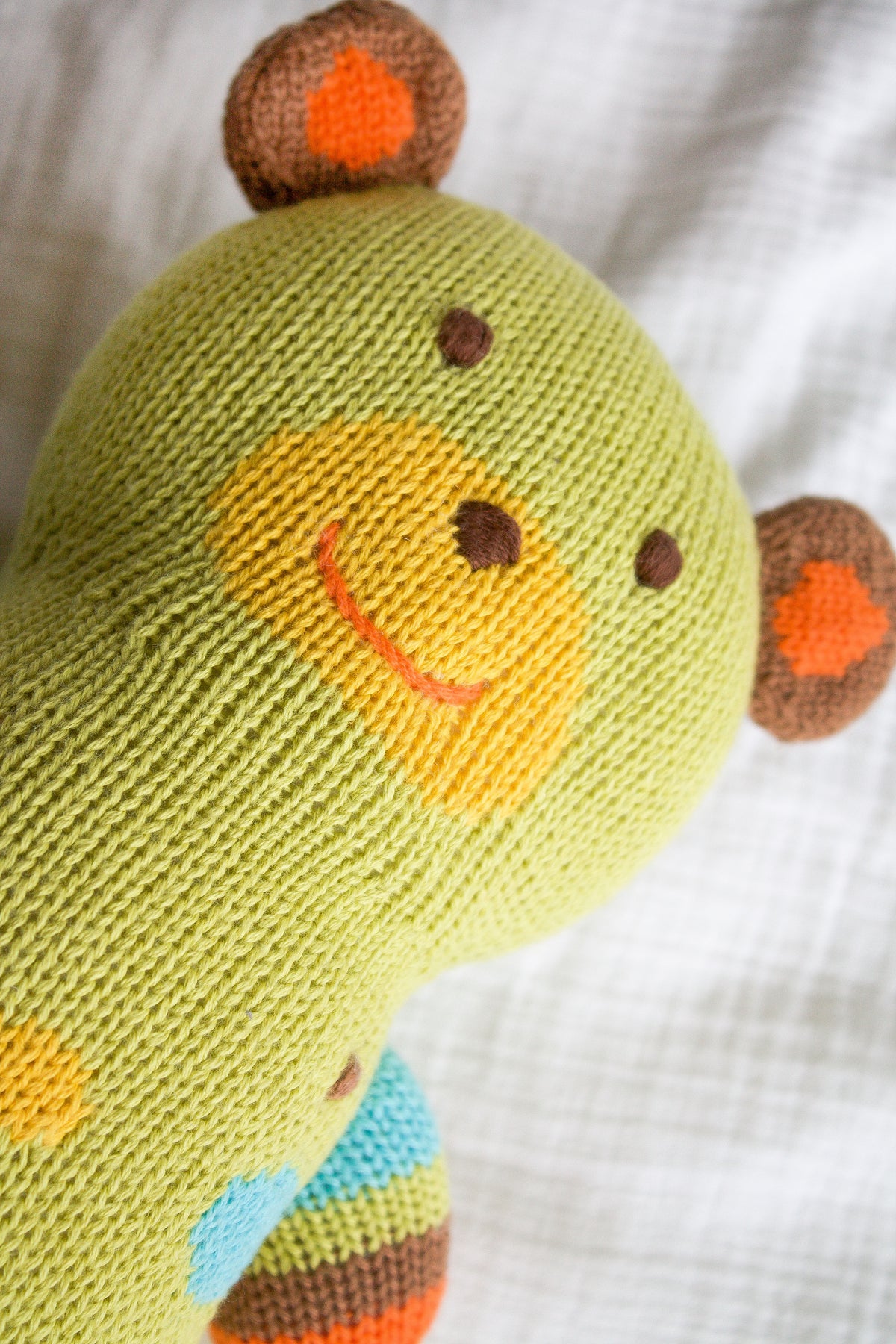 A knitted green teddy bear with a yellow snout, red smile, and brown and orange ears lies on a white fabric surface.