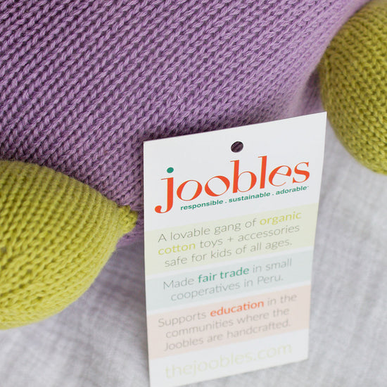 A close-up of a knitted purple and green toy with a product tag describing it as an organic, handcrafted cotton toy made through fair trade in Peru by Joobles.