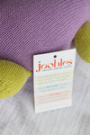 A close-up of a knitted purple and green toy with a product tag describing it as an organic, handcrafted cotton toy made through fair trade in Peru by Joobles.